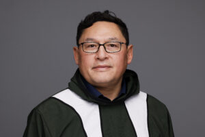 Portrait of a man in a green and white academic gown with glasses, facing the camera against a gray backdrop.